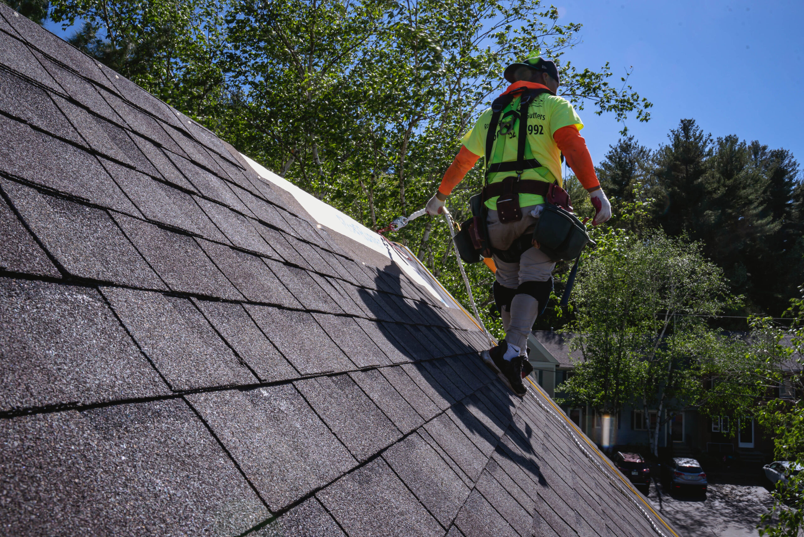 Professional roofer in safety harness and high-visibility vest working on asphalt shingle roof repair on sunny day