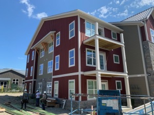 Modern three-story residential building under construction with red and gray siding, white trim, and construction equipment visible
