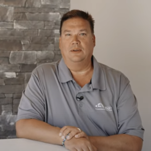 Professional man in gray polo shirt sitting at table with brick wall background, appearing to be in interview or business setting