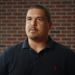Man in navy blue polo shirt with red logo standing against brick wall background, looking upward with serious expression