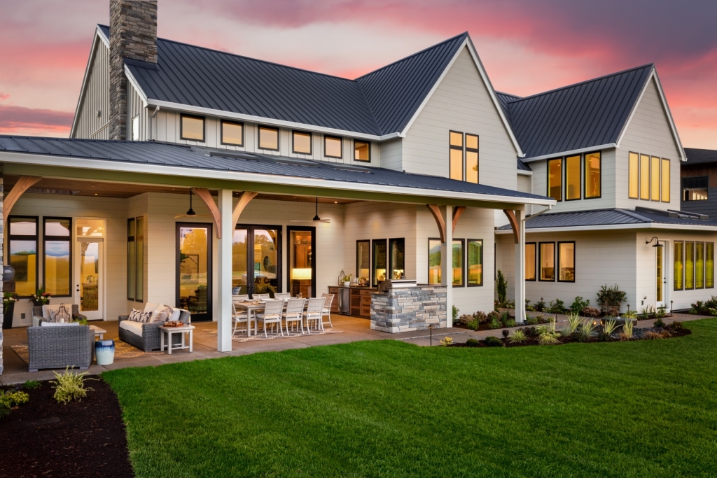 Modern farmhouse with dark metal roof, covered patio, outdoor dining area, and manicured lawn at sunset