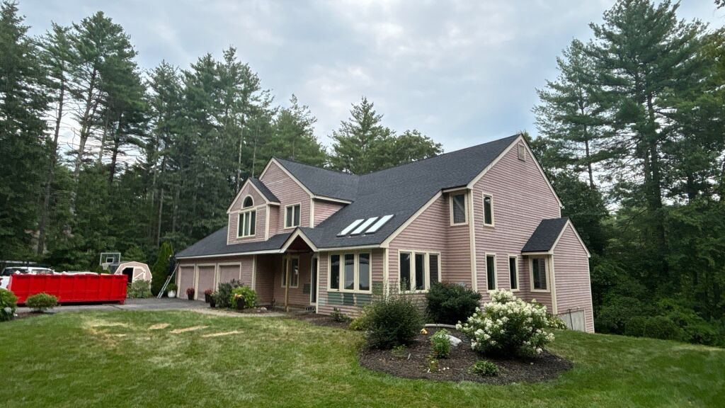 Large two-story cedar shake house with dark roof, multiple windows, skylights, and attached garage surrounded by pine trees and landscaping