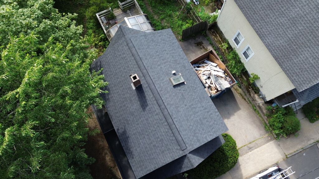Aerial view of house with new gray asphalt shingle roof, skylight, and construction dumpster filled with old roofing materials