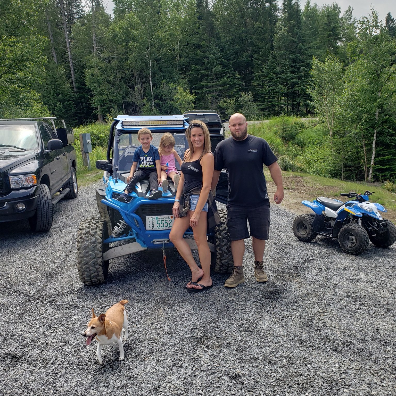 Family with two children posing by blue off-road vehicle and ATV on gravel road, with dog in foreground and forest background