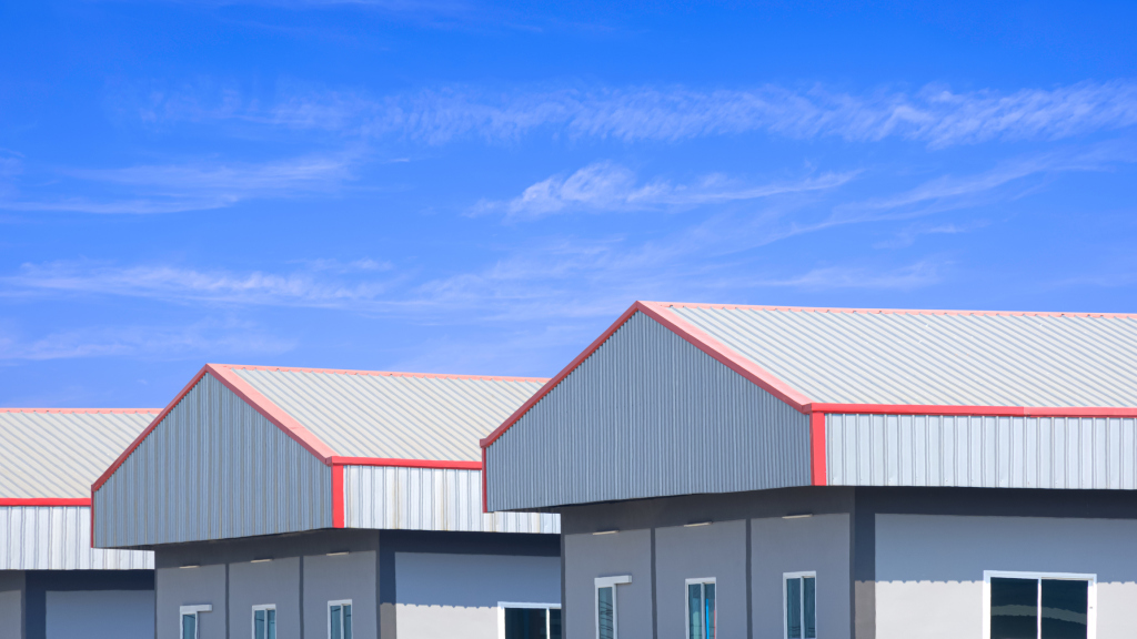Modern industrial warehouse buildings with red-trimmed metal roofing and gray walls under a bright blue sky with white clouds