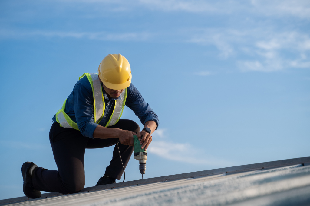 Construction worker in yellow hard hat and safety vest drilling on metal roof against blue sky