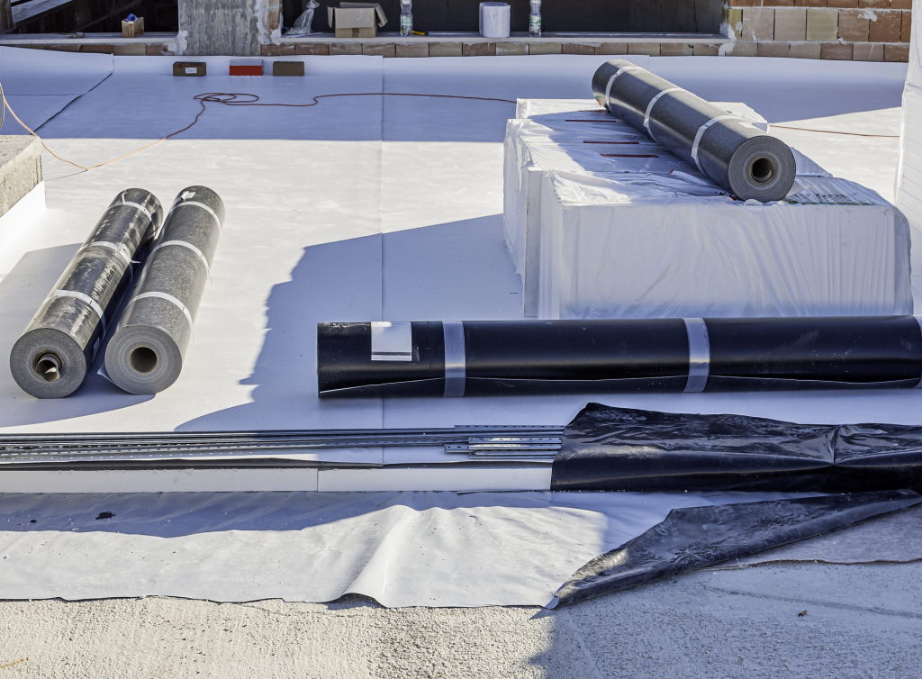 Waterproofing materials including black membrane rolls and white protective sheeting laid out on construction site for roof installation