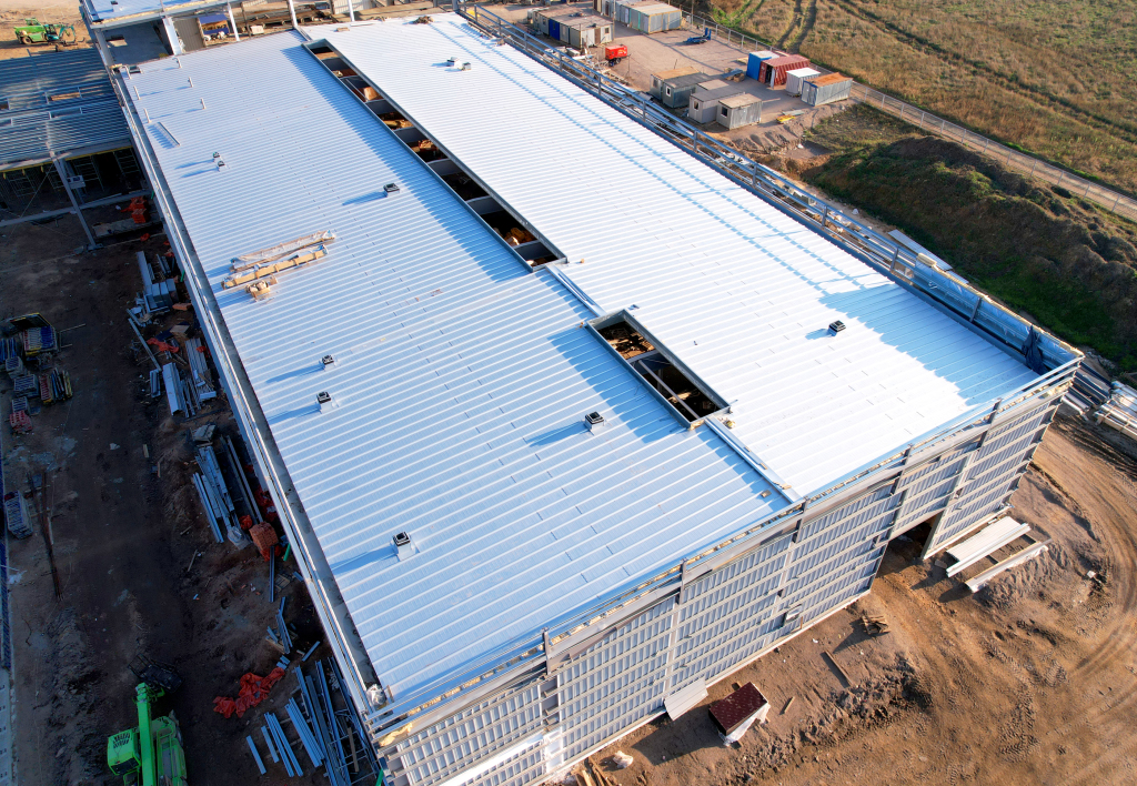 Aerial view of large industrial warehouse under construction with white metal roofing, steel framework, and construction equipment