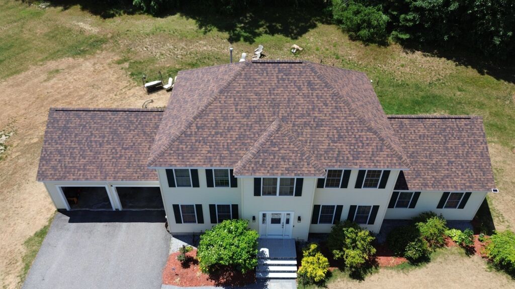 Aerial view of a large two-story colonial house with brown shingle roof, black shutters, attached garage, and landscaped yard