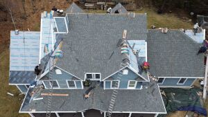 Aerial view of house roofing construction showing workers installing gray shingles with ladders and materials scattered around
