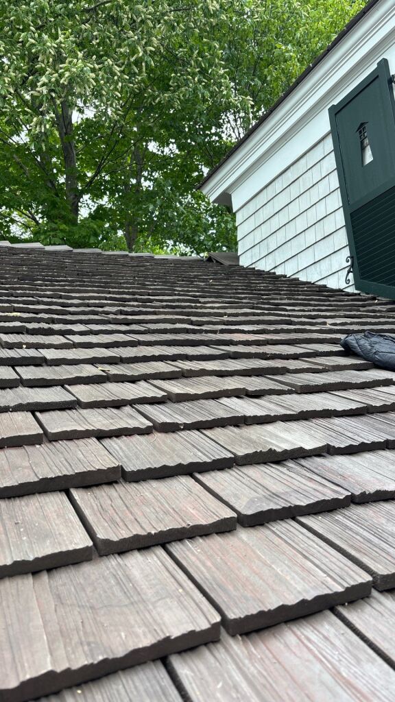 Weathered cedar shake roof shingles on residential house with green shutters and lush trees in background
