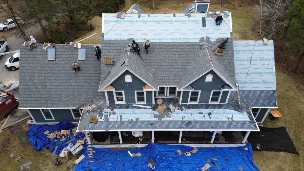 Aerial view of house under construction with workers installing gray shingles on roof, blue protective tarps and materials scattered around