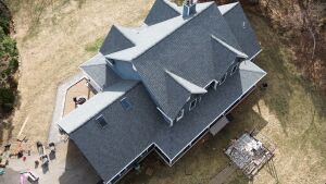 Aerial view of large residential house with dark gray shingle roof, multiple dormers, and skylights surrounded by yard and driveway