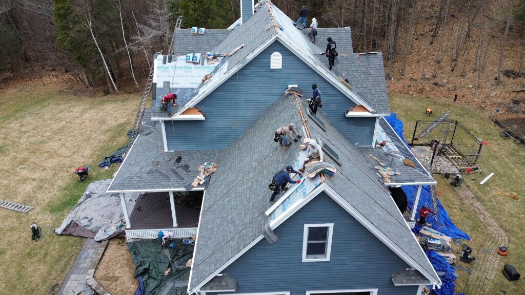 Aerial view of roofing crew installing new shingles on blue house roof with workers, equipment, and materials scattered around property