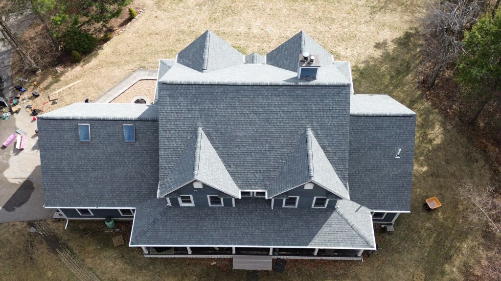 Aerial view of large house with dark gray shingle roof featuring multiple gables and dormers, surrounded by lawn and trees