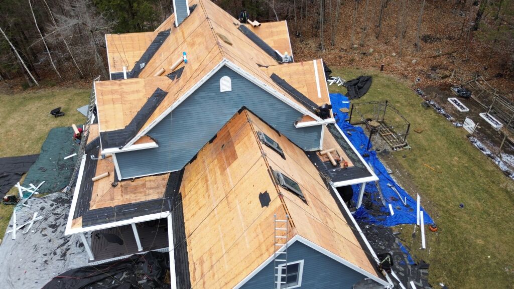 Aerial view of house under construction with exposed plywood roof decking, blue siding, and roofing materials scattered around property