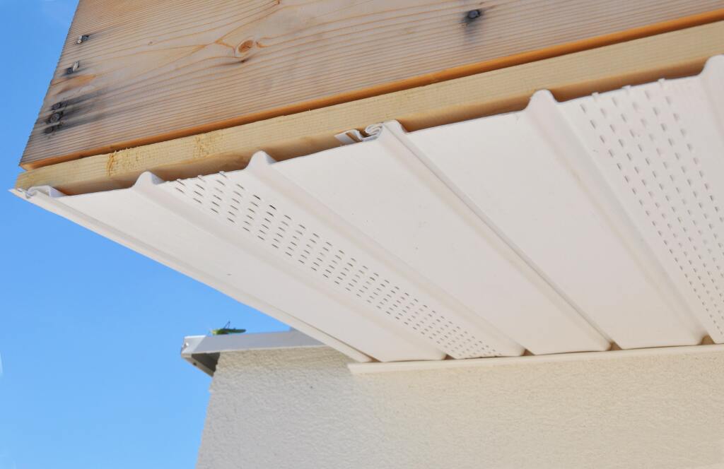 White perforated vinyl soffit panels installed under roof eaves with exposed wooden rafters against clear blue sky