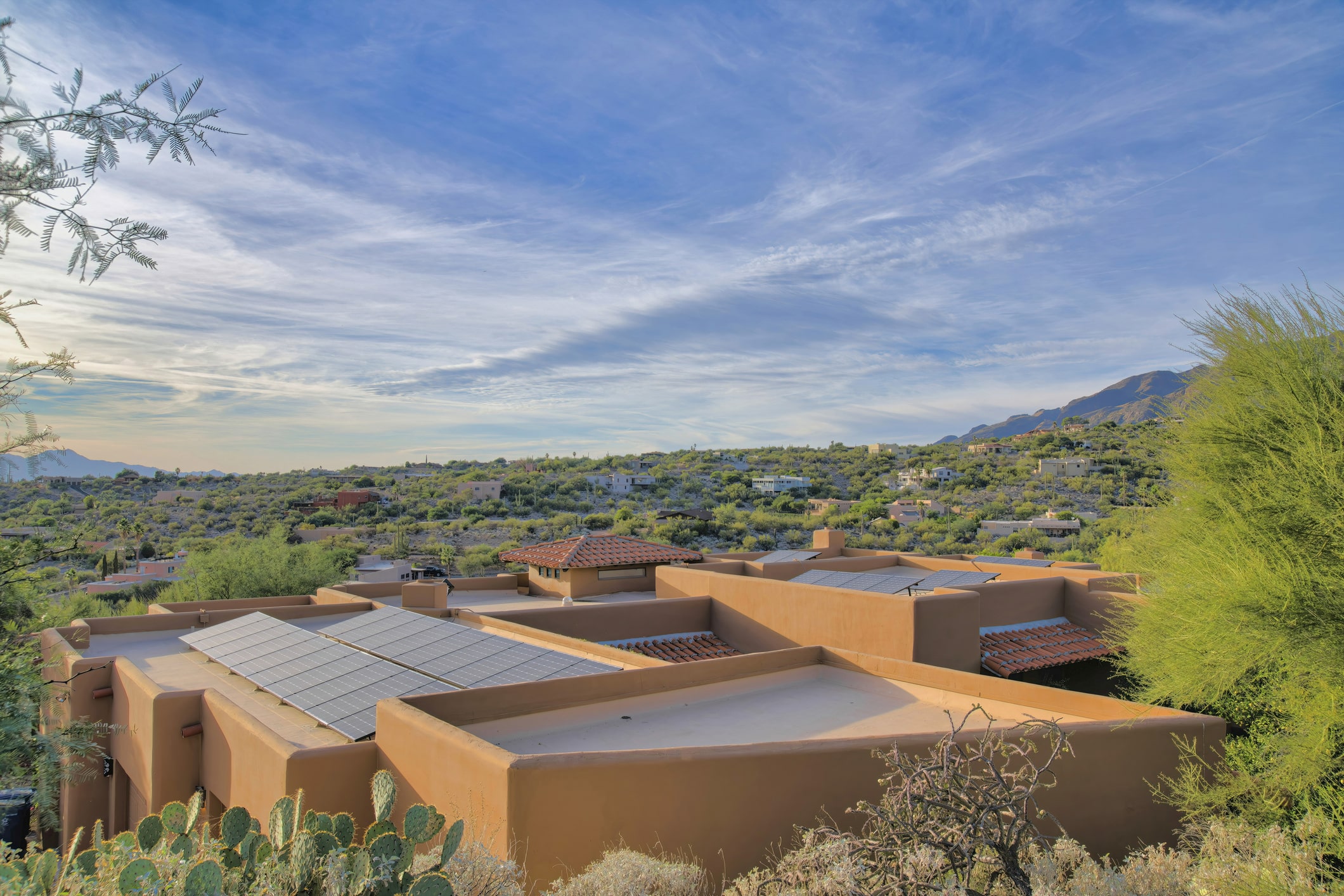 Modern adobe-style home with solar panels on flat roof, nestled in desert hills with cacti and mountain views under blue sky