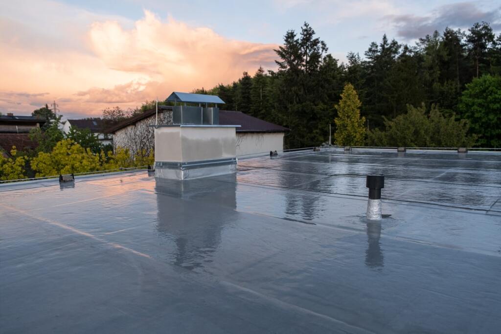 Flat commercial roof with HVAC unit and vents during sunset, showing wet membrane roofing system with residential houses in background
