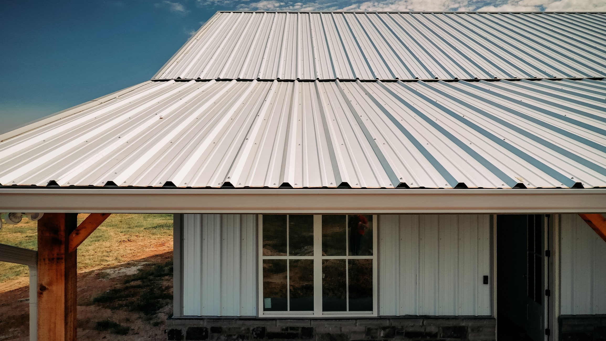 Modern farmhouse with white corrugated metal roof and siding, featuring large windows and wooden support beams under blue sky