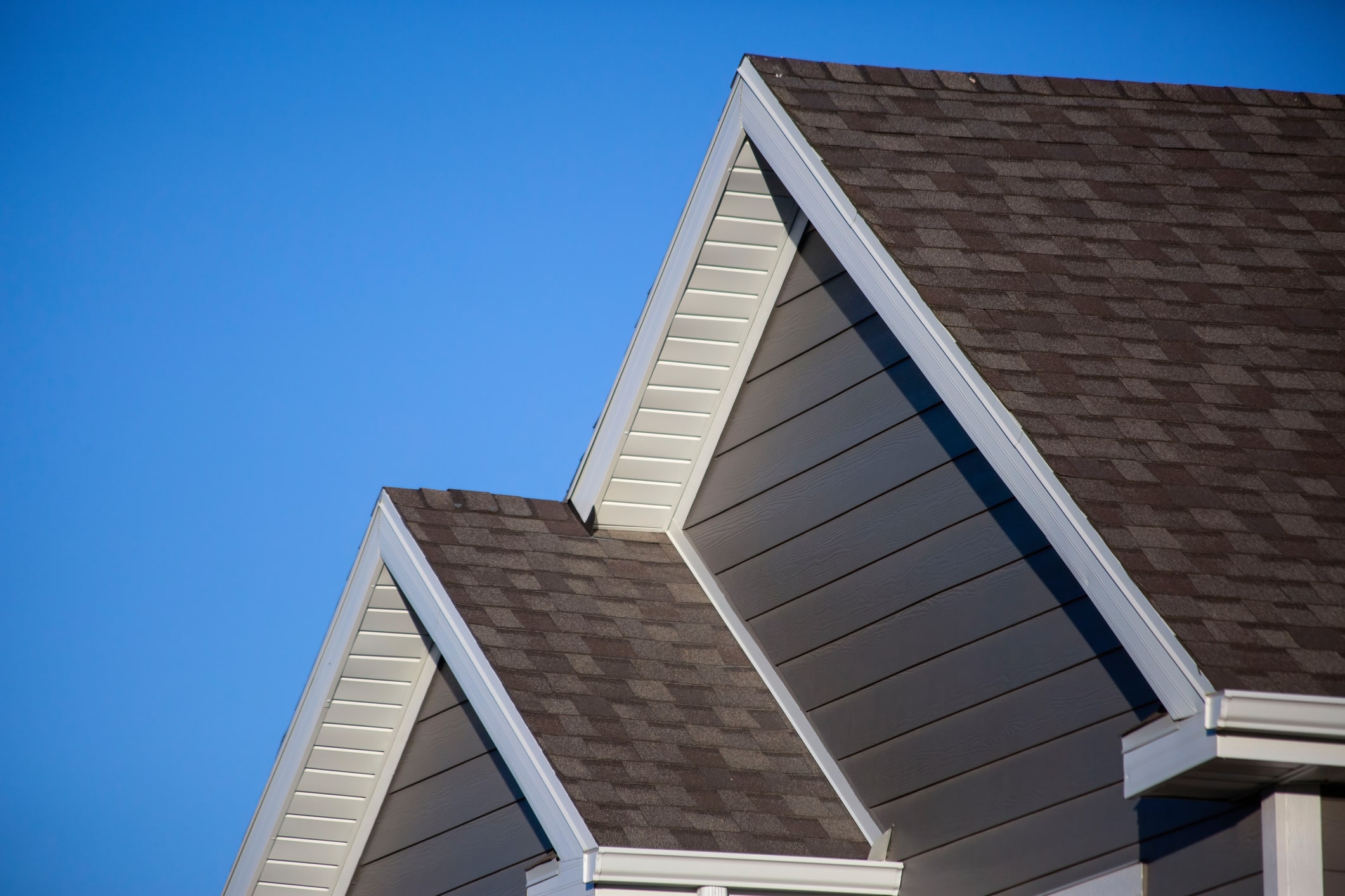 Close-up view of house roof gables with brown shingles and white trim against bright blue sky