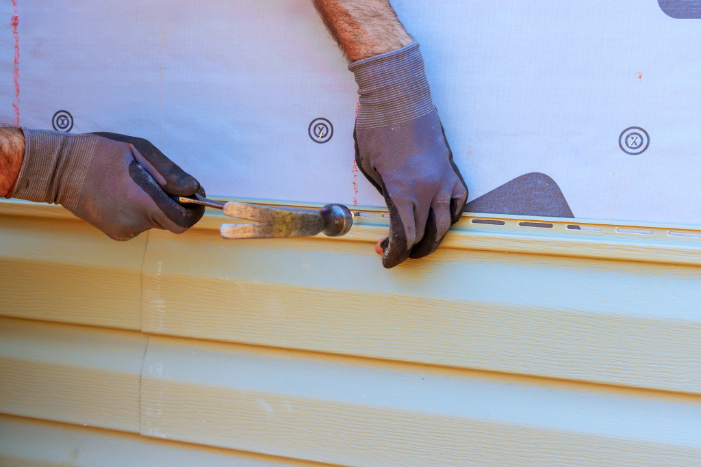 Person using wood chisel tool on light wooden board, hands wearing dark gloves for woodworking safety