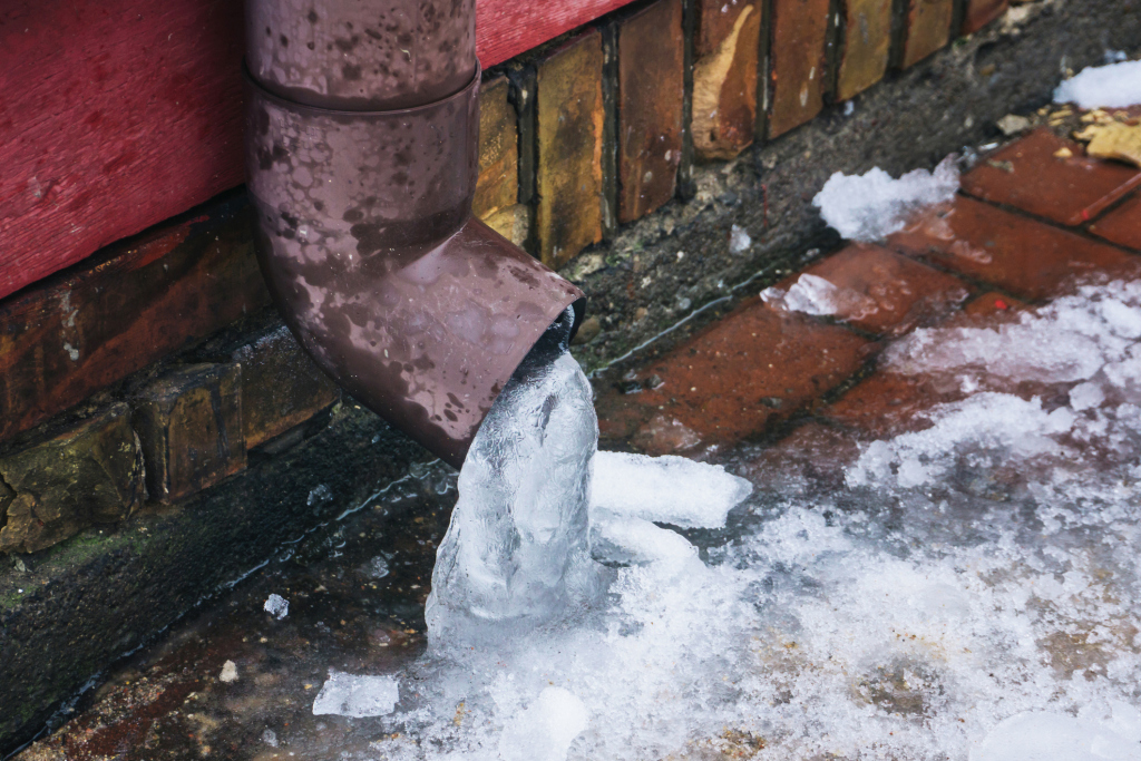 Ice formation from frozen water leak at base of brown downspout against brick wall and wet pavement