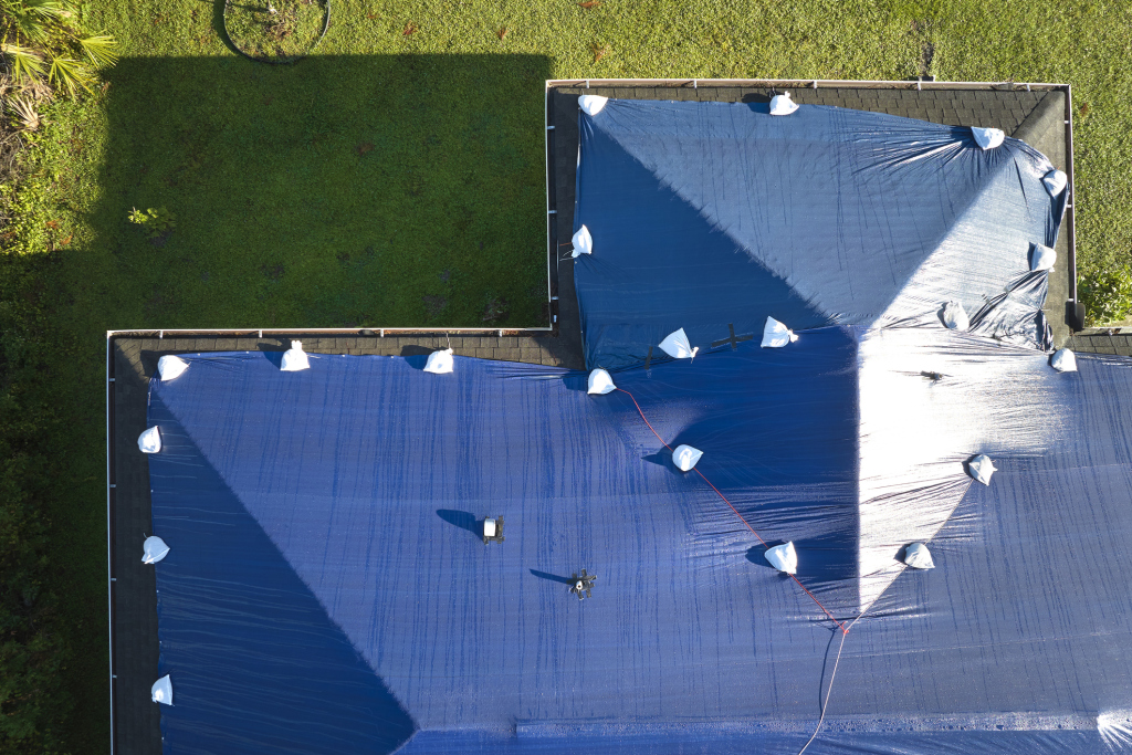 Aerial view of blue tarp covering residential roof with white sandbags securing edges, workers visible on surface during roofing project