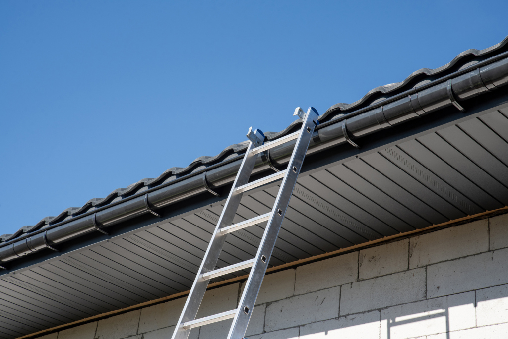 Aluminum ladder leaning against house roof with dark tiles and gutters under clear blue sky for home maintenance work