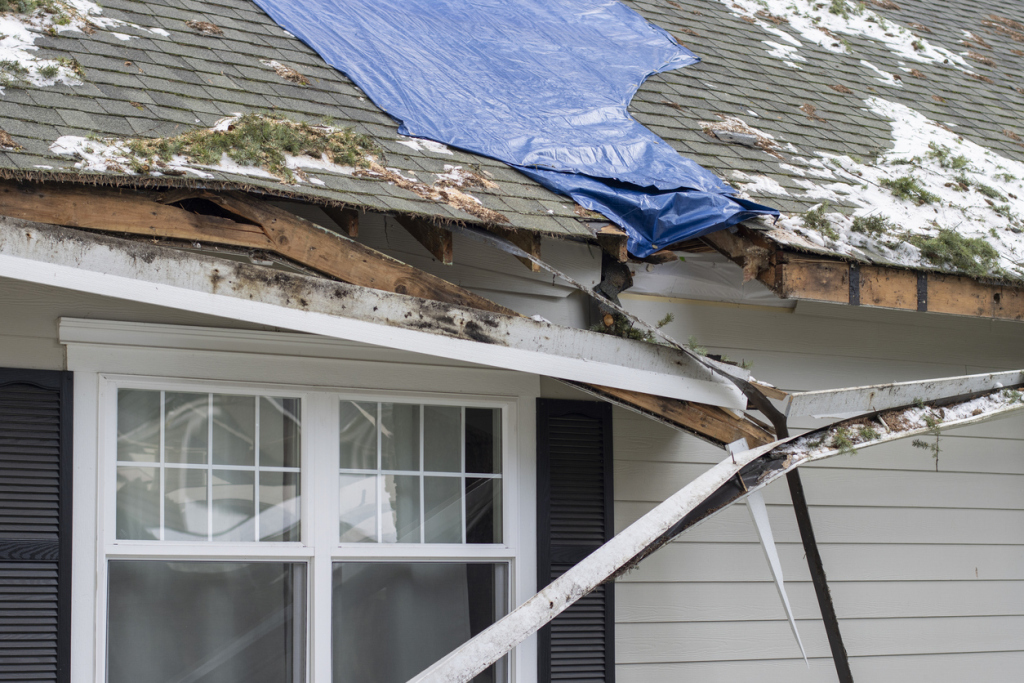 Damaged house roof with collapsed section, exposed wooden beams, blue tarp covering, and visible storm damage to shingles and gutters