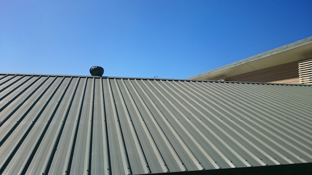 Metal corrugated roof with whirlybird ventilator against clear blue sky, showing ribbed roofing panels and building eaves