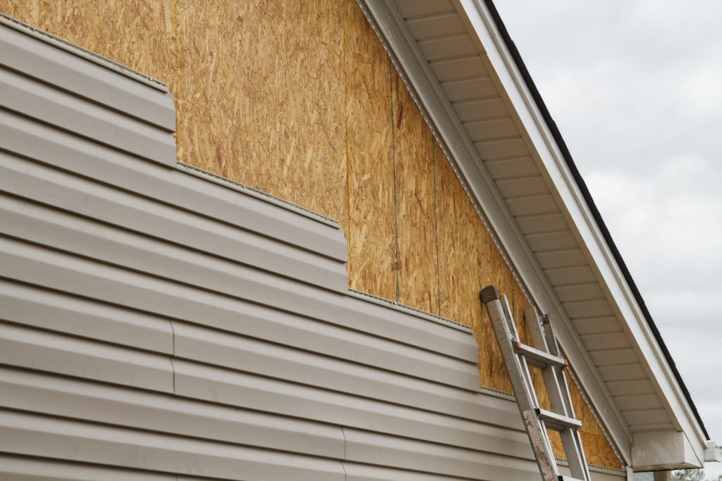 Partial home siding installation showing beige vinyl siding, exposed plywood sheathing, and aluminum ladder against house exterior