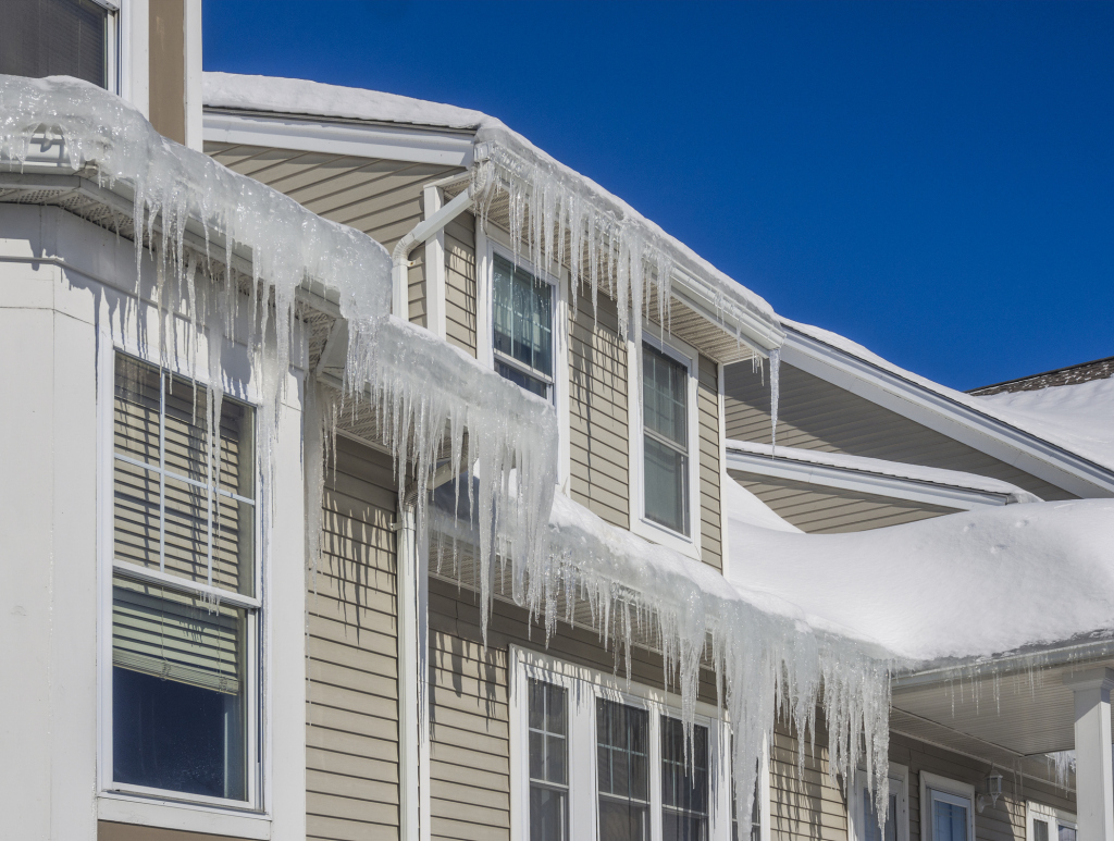 Large icicles hanging from roof and gutters of beige house with white trim against clear blue winter sky