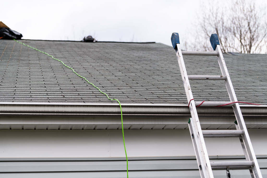 Aluminum ladder leaning against house roof with asphalt shingles, green extension cord visible, gutter cleaning or roof maintenance setup