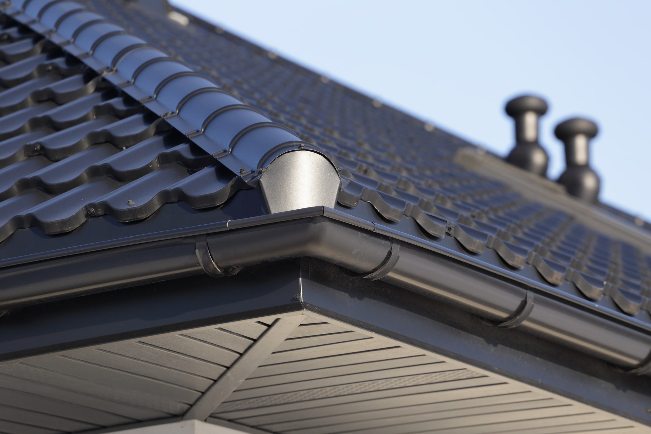 Close-up view of dark tile roof with metal gutter system and chimney stacks against blue sky