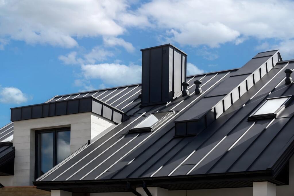 Modern gray metal roof with standing seam panels, skylights, chimney and white dormer window against blue cloudy sky