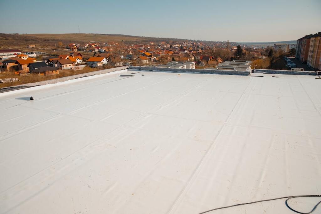 White flat roof membrane covering large commercial building with residential town and orange-tiled houses visible in background