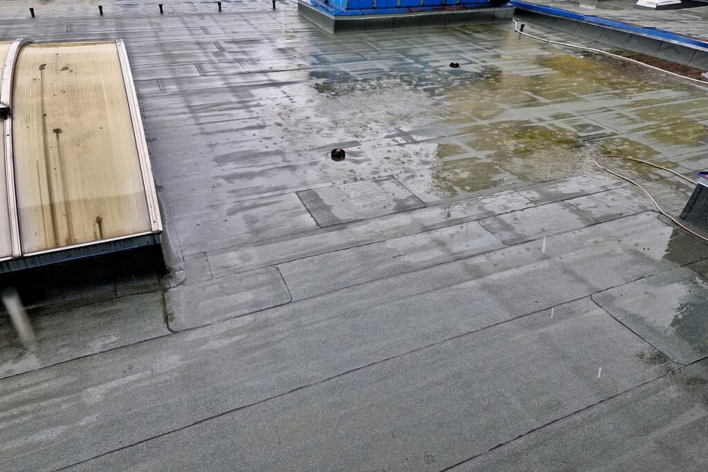 Flat commercial roof with wet membrane surface, algae growth, and construction materials including plywood and blue equipment