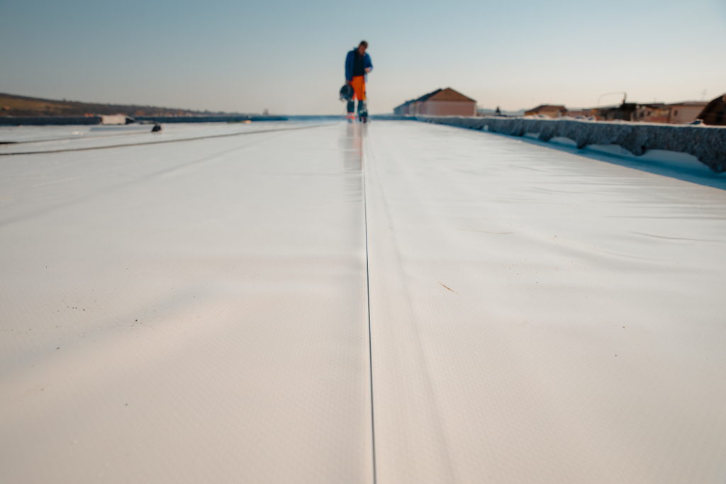 Worker in blue shirt and orange pants standing on white rooftop surface with buildings in background during daytime