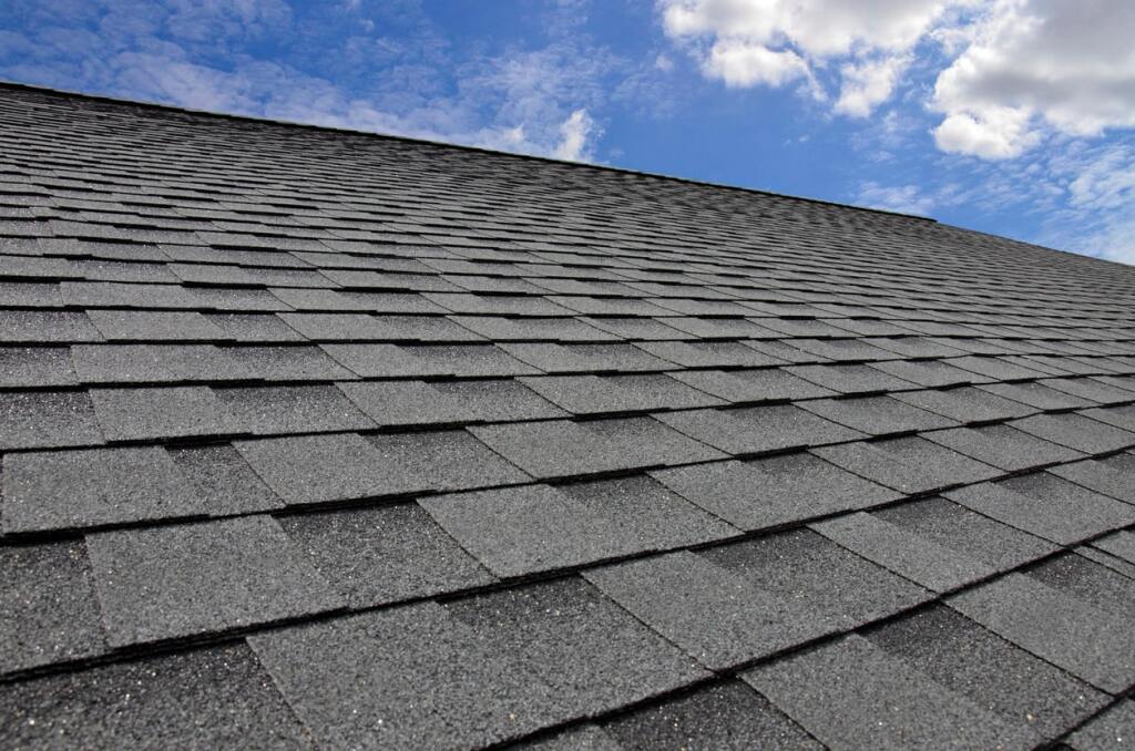 Gray asphalt shingle roof photographed from below against blue cloudy sky, showing overlapping rectangular shingles in rows