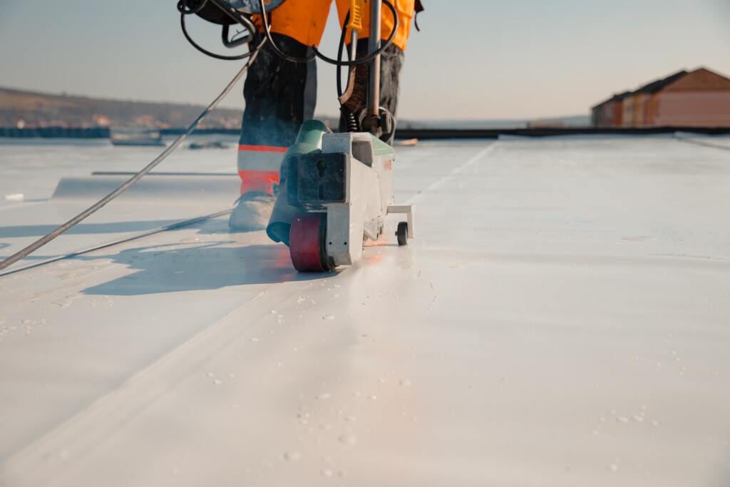 Worker in orange safety vest using industrial roofing equipment on white membrane roof surface during installation or repair work